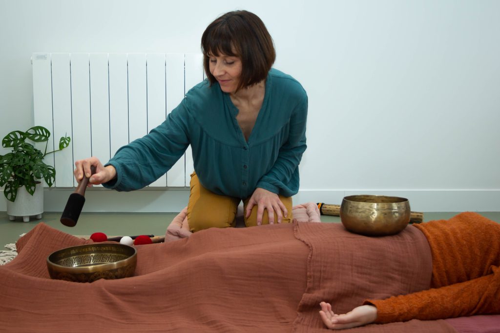 Émilie qui pratique le massage sonore aux bols tibétains. Elle se tient à genoux sur le côté droit de la patiente. La patiente est allongée sur le dos avec une fine couverture sur le bas du corps