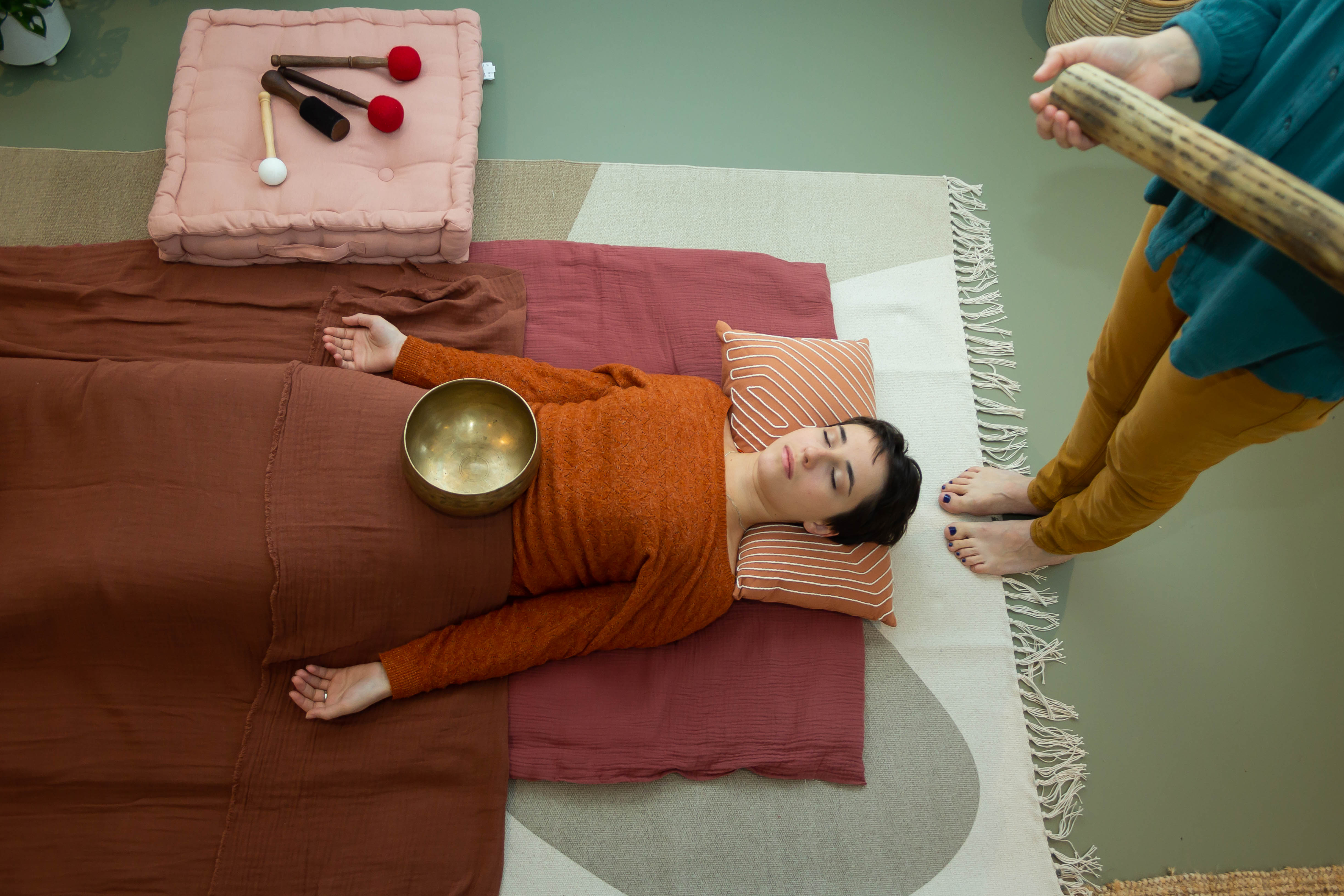 Une patiente est allongée sur le dos, sur un tapis mauve avec un coussin orange et blanc. Elle à les bras tendus et un bols est posé sur son abdomen. Émilie est pieds nu, et se tient debout derrière la patiente avec un bâton