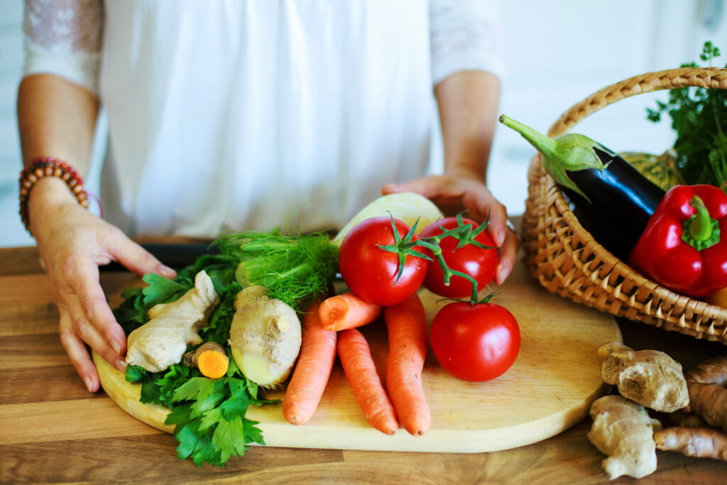 Une personne se trouve devant une planche de divers légumes: tomates, carottes, gingembre, persil. À côté se trouve un panier contenant un poivron et une aubergine