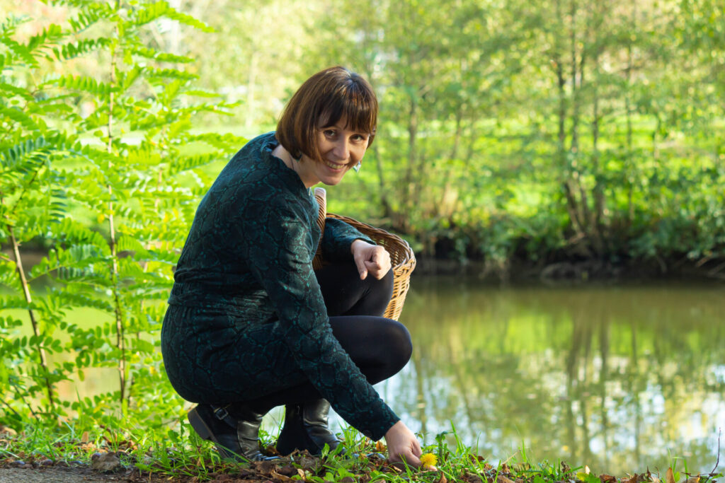 Émilie sourit, accroupie au bord de l'eau d'un étang dans la nature, avec un panier en osier sous le bras